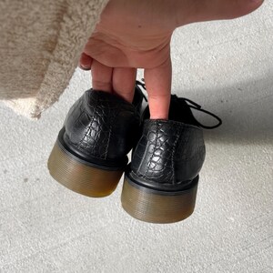 Puede incluir: Zapatos de cuero negro con un patr&oacute;n texturizado similar al de un cocodrilo. Los zapatos tienen suelas gruesas de color marr&oacute;n y cordones negros. Los zapatos est&aacute;n siendo sostenidos por una mano.