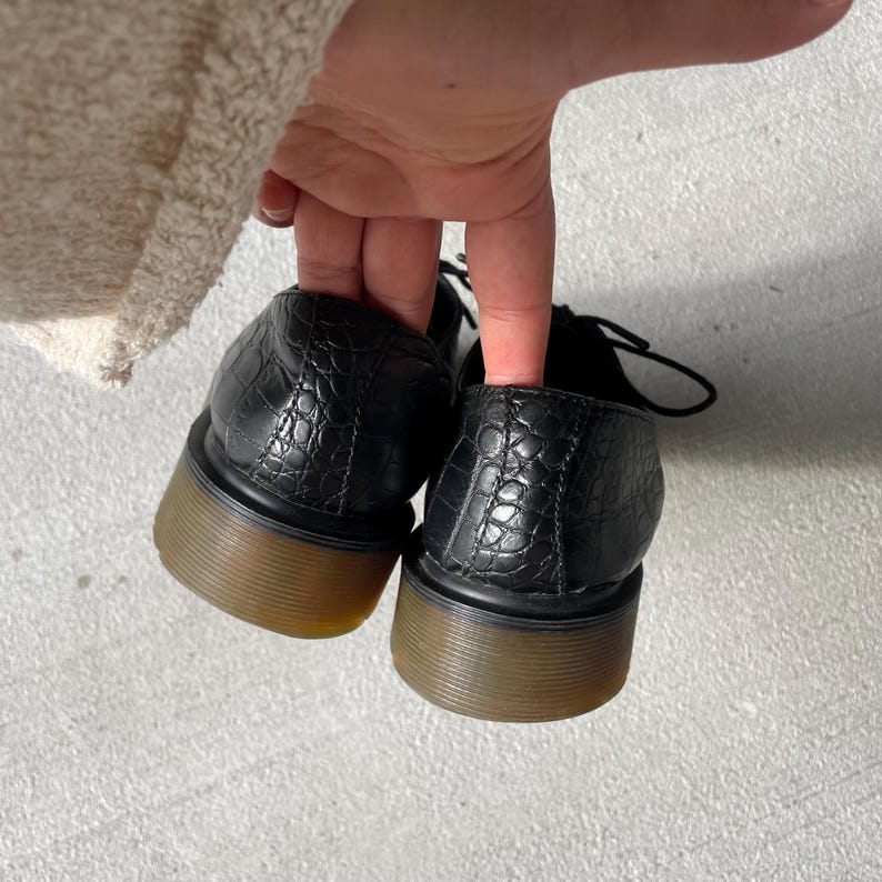 Puede incluir: Zapatos de cuero negro con un patr&oacute;n texturizado similar al de un cocodrilo. Los zapatos tienen suelas gruesas de color marr&oacute;n y cordones negros. Los zapatos est&aacute;n siendo sostenidos por una mano.