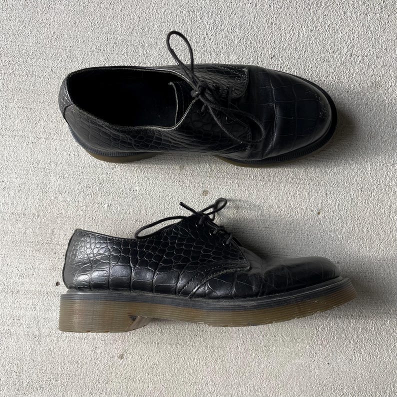 Puede incluir: Par de zapatos de cuero negro con textura de piel de cocodrilo. Los zapatos tienen un dise&ntilde;o con cordones y una suela gruesa de color marr&oacute;n claro. Los zapatos se muestran sobre una superficie gris claro.