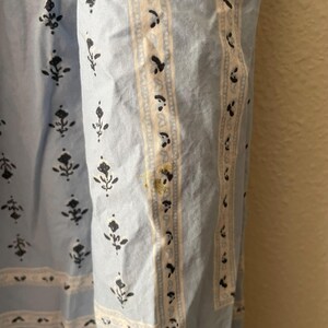 Pu&ograve; includere: Primo piano di un tessuto azzurro con motivo floreale. Il tessuto presenta un motivo ripetuto di motivi floreali neri e bordi in pizzo bianco. Una striscia verticale con sfondo color crema e accenti neri corre lungo il lato. Ci sono macchie gialle sul tessuto.
