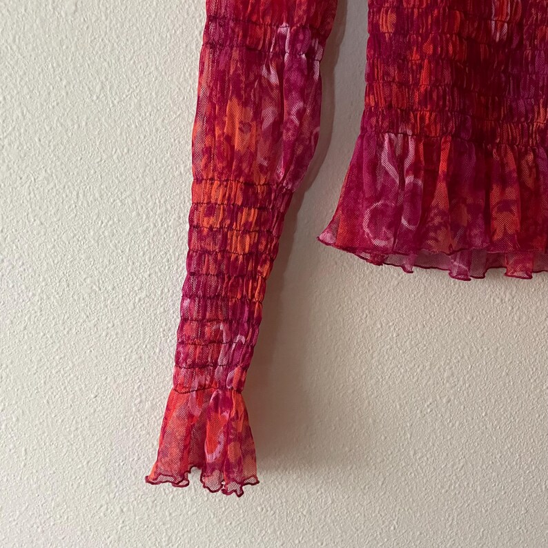 May include: A close-up of a sheer, long-sleeved top in shades of red and orange. The fabric has a tie-dye effect, with a gathered bodice and ruffled cuffs. The top is made of a lightweight, semi-transparent material.
