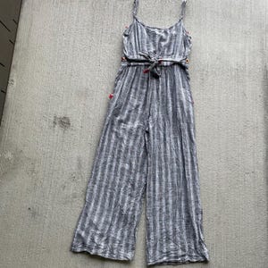 K&ouml;nnte beinhalten: Ein grau-wei&szlig; gestreifter Jumpsuit mit Spaghettitr&auml;gern und einem Bindeg&uuml;rtel. Der Jumpsuit hat weite Beine und rote Akzente an den Seiten und an der Taille. Der Stoff scheint leicht zu sein.