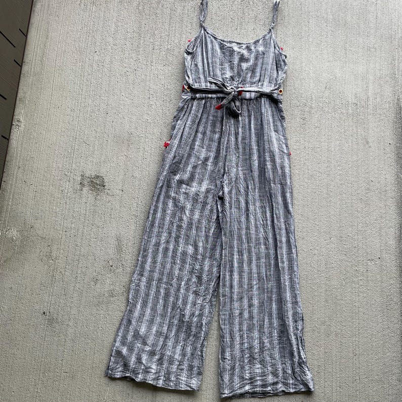 K&ouml;nnte beinhalten: Ein grau-wei&szlig; gestreifter Jumpsuit mit Spaghettitr&auml;gern und einem Bindeg&uuml;rtel. Der Jumpsuit hat weite Beine und rote Akzente an den Seiten und an der Taille. Der Stoff scheint leicht zu sein.
