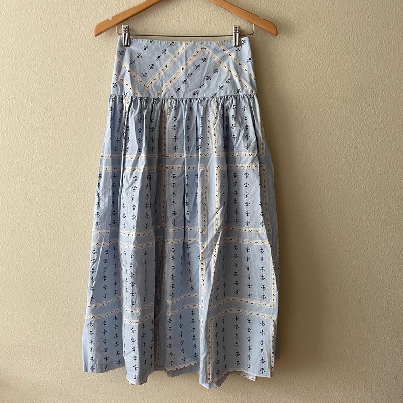 Pu&ograve; includere: Gonna azzurra con un motivo bianco e blu navy, appesa a una gruccia di legno. La gonna presenta una vita alta e un design fluido di media lunghezza. Il motivo include piccoli elementi floreali e geometrici.