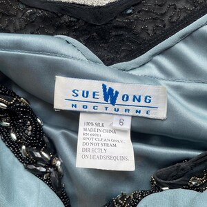 K&ouml;nnte beinhalten: Ein hellblaues Seidenkleid mit schwarzen Perlenbesatz. Das Kleid tr&auml;gt ein Sue Wong Nocturne-Etikett mit Pflegehinweisen: 100 % Seide, hergestellt in China, RN 69701, nur chemische Reinigung, nicht direkt auf Perlen/Pailletten dampfen.