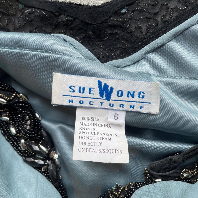 K&ouml;nnte beinhalten: Ein hellblaues Seidenkleid mit schwarzen Perlenbesatz. Das Kleid tr&auml;gt ein Sue Wong Nocturne-Etikett mit Pflegehinweisen: 100 % Seide, hergestellt in China, RN 69701, nur chemische Reinigung, nicht direkt auf Perlen/Pailletten dampfen.
