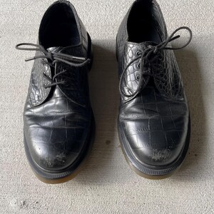 Puede incluir: Par de zapatos de vestir de cuero negro con un patr&oacute;n texturizado similar al de un cocodrilo. Los zapatos tienen un dise&ntilde;o cl&aacute;sico con cordones y una puntera redondeada. Las suelas son de color marr&oacute;n claro en contraste. Los zapatos muestran signos de desgaste.