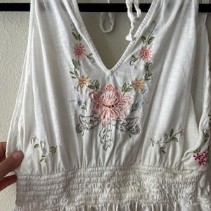 May include: White halter top with a V-neckline and floral embroidery in pink, yellow, green, and gray. The top features a shirred waist and a tie closure at the neck. The embroidery is centered on the chest and along the neckline.