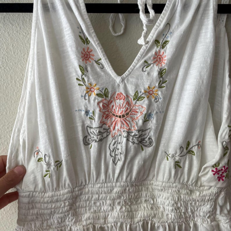 May include: White halter top with a V-neckline and floral embroidery in pink, yellow, green, and gray. The top features a shirred waist and a tie closure at the neck. The embroidery is centered on the chest and along the neckline.