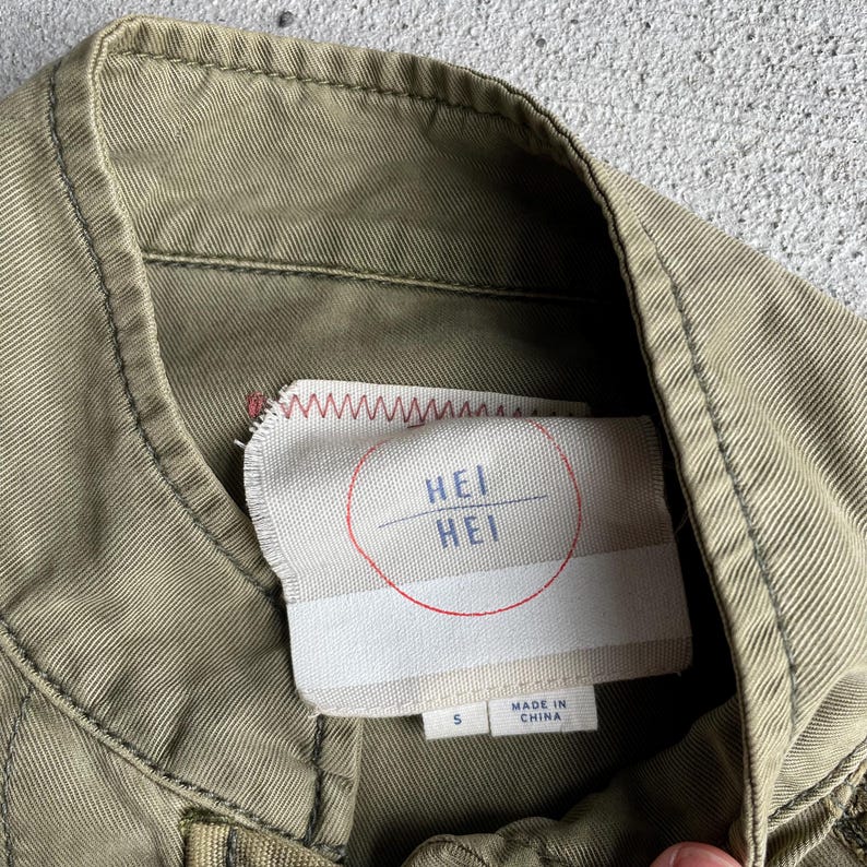 Pode incluir: Close-up de uma gola de jaqueta verde-oliva com uma etiqueta costurada. A etiqueta apresenta o texto "HEI HEI" dentro de um c&iacute;rculo vermelho, com "MADE IN CHINA" impresso abaixo. A jaqueta &eacute; tamanho P.