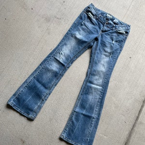 Puede incluir: Un par de pantalones vaqueros de mezclilla azul claro con detalles desgastados. Los pantalones tienen un estilo de pierna acampanada y están colocados sobre una superficie de concreto gris.