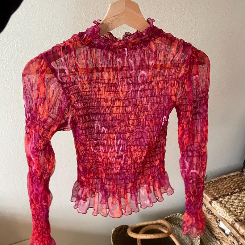 May include: A sheer, long-sleeved top in shades of red and orange, hanging on a wooden hanger. The top features a high neckline, a ruched bodice, and a ruffled hemline. The fabric has a subtle, patterned design.