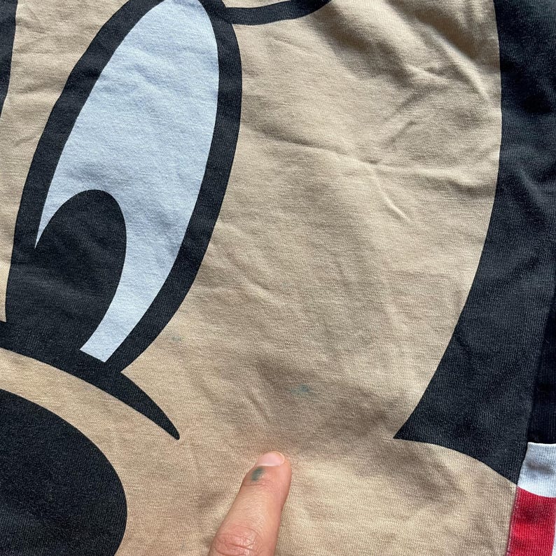 May include: Close-up of a tan fabric featuring a cartoon character's face. The character's eye is white with a black outline, and the face has black and tan sections. A finger is touching the fabric.