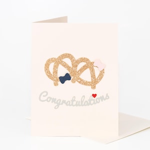 Op de afbeelding: Een gevouwen crème-kleurige wenskaart met een goudglitter pretzel ontwerp, een marineblauwe vlinderdas en een roze strik. Het woord "Gefeliciteerd" is in zilveren letters geschreven, met een klein rood hartje eronder.