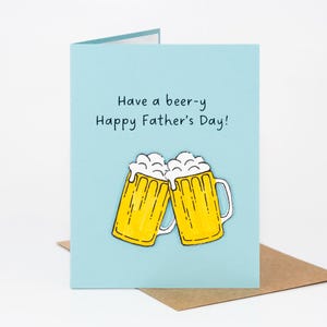 以下が含まれることがあります： 「Have a beer-y Happy Father's Day!」のテキストと2つの漫画のビールジョッキが描かれた、水色の父の日カード。ビールジョッキは黄色で、白い泡と白いハンドルが付いています。カードは茶色の封筒の上にあります。