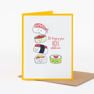 Puede incluir: Una tarjeta de felicitación con borde amarillo y fondo blanco presenta ilustraciones de sushi de dibujos animados. El texto "So happy you ROLL with me" está escrito en rojo. La tarjeta está apoyada en un sobre marrón.