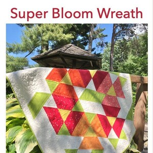 May include: A quilt with a geometric pattern of red, orange, yellow, and green triangles. The quilt is hanging on a wooden railing in a garden setting. The text "Super Bloom Wreath" is at the top of the image. The text "pattern by Jill Blair" is at the bottom left of the image. The text "materiella.com" is at the bottom right of the image.