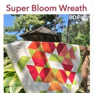 May include: A quilt pattern called "Super Bloom Wreath" by Jill Blair. The quilt is made of red, orange, yellow, and green fabric. The quilt is hanging on a wooden railing in a garden setting.