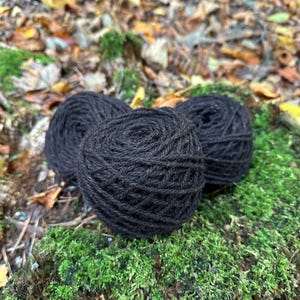 May include: Three black yarn balls sit on green moss. The yarn is tightly wound, showing the texture of the fibres. The background has fallen leaves, suggesting an outdoor setting. Ideal for knitting or crochet projects, the yarn is a deep, rich black.