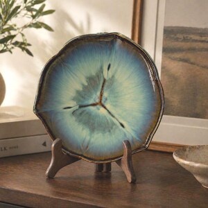 May include: A decorative ceramic plate with a unique, organic shape, displayed on a wooden stand. The plate features a blue and brown glaze with a radial pattern, resembling a cross-section of a tree trunk. The plate is approximately 10 inches in diameter.