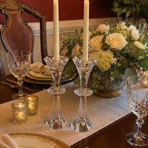 May include: Two lit ivory taper candles in elegant crystal candlesticks. The candlesticks have a detailed, faceted design. The table setting includes crystal glassware, gold-rimmed plates, and a floral centerpiece, creating a sophisticated dining scene.