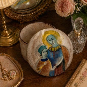 Peut inclure: Boîte à bijoux en céramique avec couvercle orné d'une illustration peinte de deux personnages aux accents bleus et jaunes. La boîte est ouverte, révélant un intérieur clair. Une lampe et un miroir dorés sont en arrière-plan.
