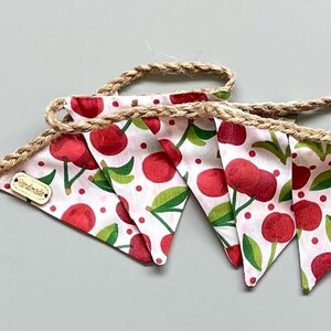 May include: A handmade fabric bunting featuring a cherry pattern. White fabric triangles display red cherries, green leaves, and red dots. The bunting is strung on a natural jute rope. The tag reads "handmade".