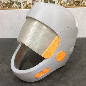 May include: A white helmet with an orange visor and orange buttons. The helmet has a futuristic design.