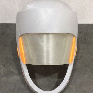 May include: A white helmet with a clear visor and orange accents. The helmet has a rounded top and a curved chin strap.