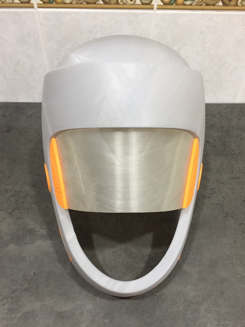 May include: A white helmet with a clear visor and orange accents. The helmet has a rounded top and a curved chin strap.