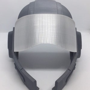May include: A gray 3D printed helmet with a clear visor. The helmet has a futuristic design with a rounded top and a curved visor.