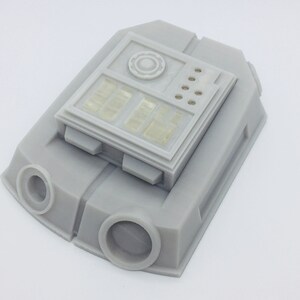 May include: A gray 3D printed model of a small, rectangular device with a panel of buttons and a circular dial. The device has a textured surface and is mounted on a base.