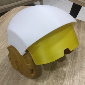 May include: A white and yellow helmet with a rounded top and a gold-colored base. The helmet has a visor that is partially down.
