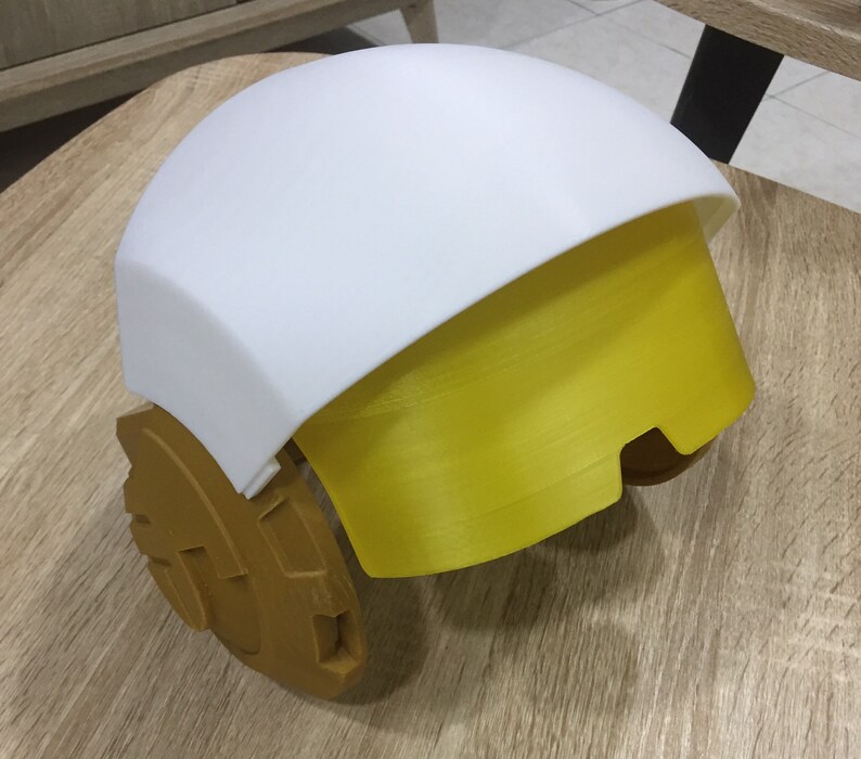 May include: A white and yellow helmet with a rounded top and a gold-colored base. The helmet has a visor that is partially down.