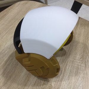 May include: A white and gold 3D printed helmet with a black visor. The helmet has a rounded shape and a textured surface.