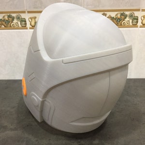 May include: A white 3D printed helmet with an orange detail on the side. The helmet has a smooth, rounded surface and a slightly angled top.
