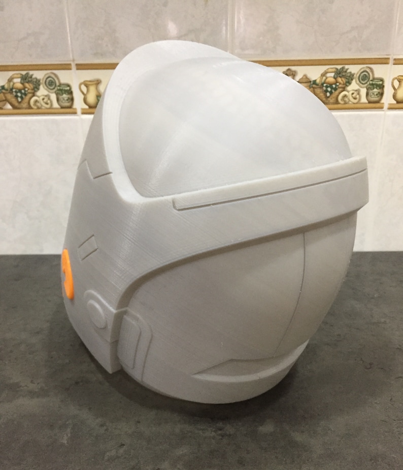 May include: A white 3D printed helmet with an orange detail on the side. The helmet has a smooth, rounded surface and a slightly angled top.
