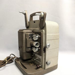May include: Vintage beige and brown film projector with a handle and various knobs. The projector has a rectangular base and a power cord. The brand name "Bell & Howell" is visible on the side.