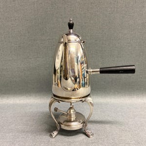 May include: A polished silver coffee pot with a black handle and a decorative black finial. The pot sits on a silver stand with ornate legs and a burner. The coffee pot is a vintage item.