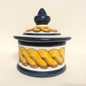 May include: A white ceramic jar with a blue and yellow rope design. The jar has a lid with a blue knob.
