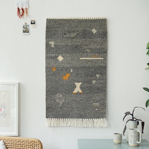 Op de afbeelding: Een grijze geweven wandhanger met geometrische patronen in wit, oranje en zwart. De hanger heeft een franje rand.