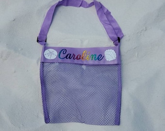 May include: Purple mesh beach bag with a white strap and a name tag that says "Caroline" in rainbow glitter letters. The tag also has a seashell and a starfish design.