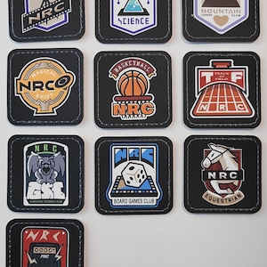 May include: A collection of ten square black patches with rounded corners and grey stitched borders. Each patch features a unique graphic design representing different clubs, including Film Research, Science, Mountain Lovers, Basketball, and Track and Field.