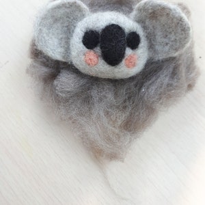 May include: A handmade felted koala bear with gray fur and large, rounded ears. The koala's face is light gray with a black nose and two small, pink cheeks. The koala is made of soft, fluffy wool.