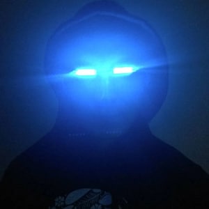 May include: A person wearing a dark hooded sweatshirt with two bright blue lights shining from the eyes.