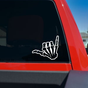 May include: White decal of a skeleton hand making the shaka sign. The sticker is applied to the rear window of a red truck. The shaka hand gesture is a symbol of friendship and solidarity.