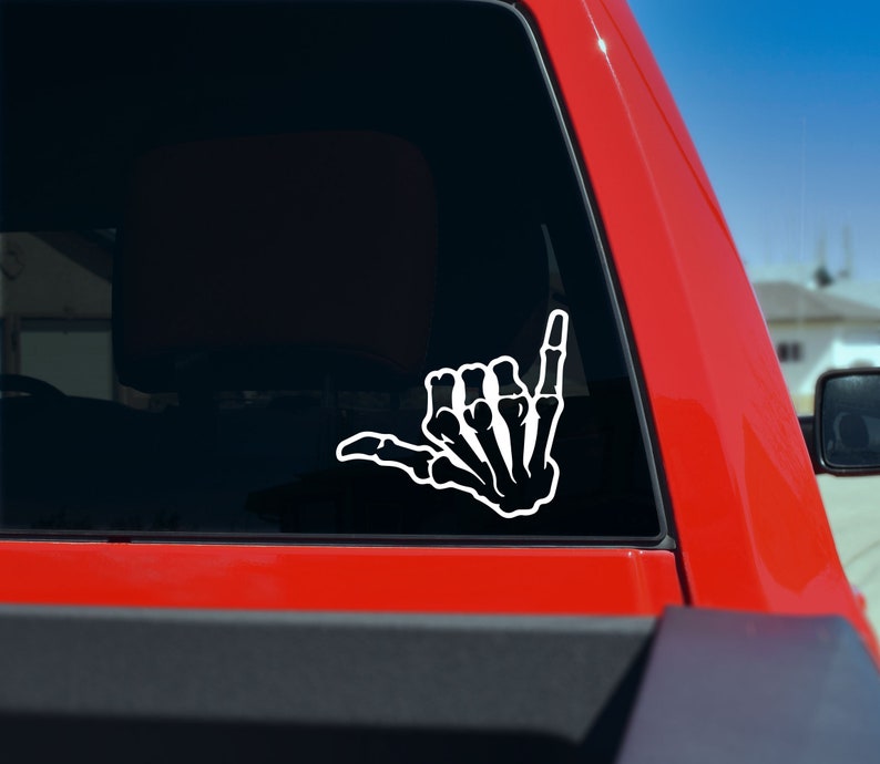 May include: White decal of a skeleton hand making the shaka sign. The sticker is applied to the rear window of a red truck. The shaka hand gesture is a symbol of friendship and solidarity.
