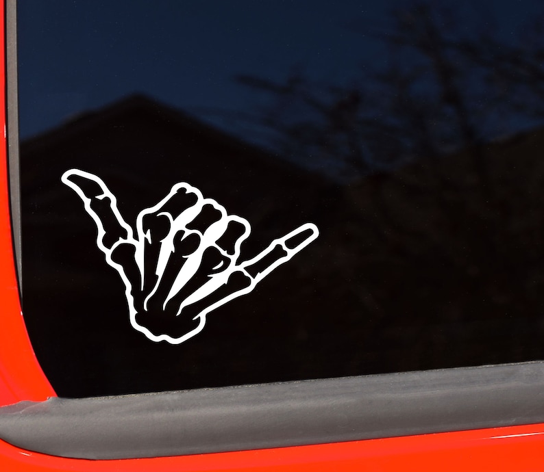 May include: White skeleton hand in the shaka gesture, a symbol of surfing culture, is displayed on a dark background. The sticker is likely for a car window or similar surface. The design is a bold, graphic representation.