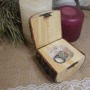 May include: A rustic wooden ring box with a bark exterior and a hinged lid. The box is open and contains two wedding bands on a white lace cushion. The box is sitting on a burlap surface with a white lace trim.
