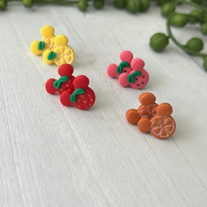 May include: Four pairs of colorful fruit-shaped earrings. Each pair is shaped like Mickey Mouse ears with a different fruit design: lemon, strawberry, orange, and watermelon.